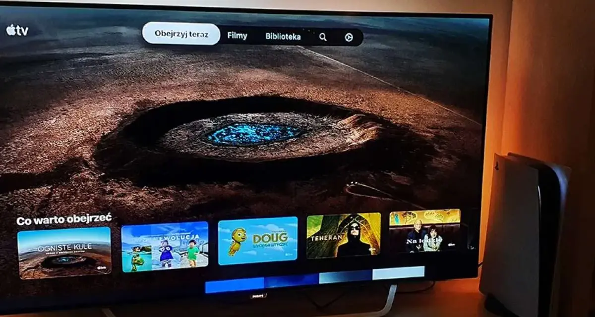Telewizor z interfejsem Apple TV i konsola PS5 obok. Na ekranie widać menu z filmami i serialami, np. "Ogniste Kule" i "Moja Ewolucja".