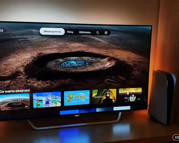 Telewizor z interfejsem Apple TV i konsola PS5 obok. Na ekranie widać menu z filmami i serialami, np. "Ogniste Kule" i "Moja Ewolucja".